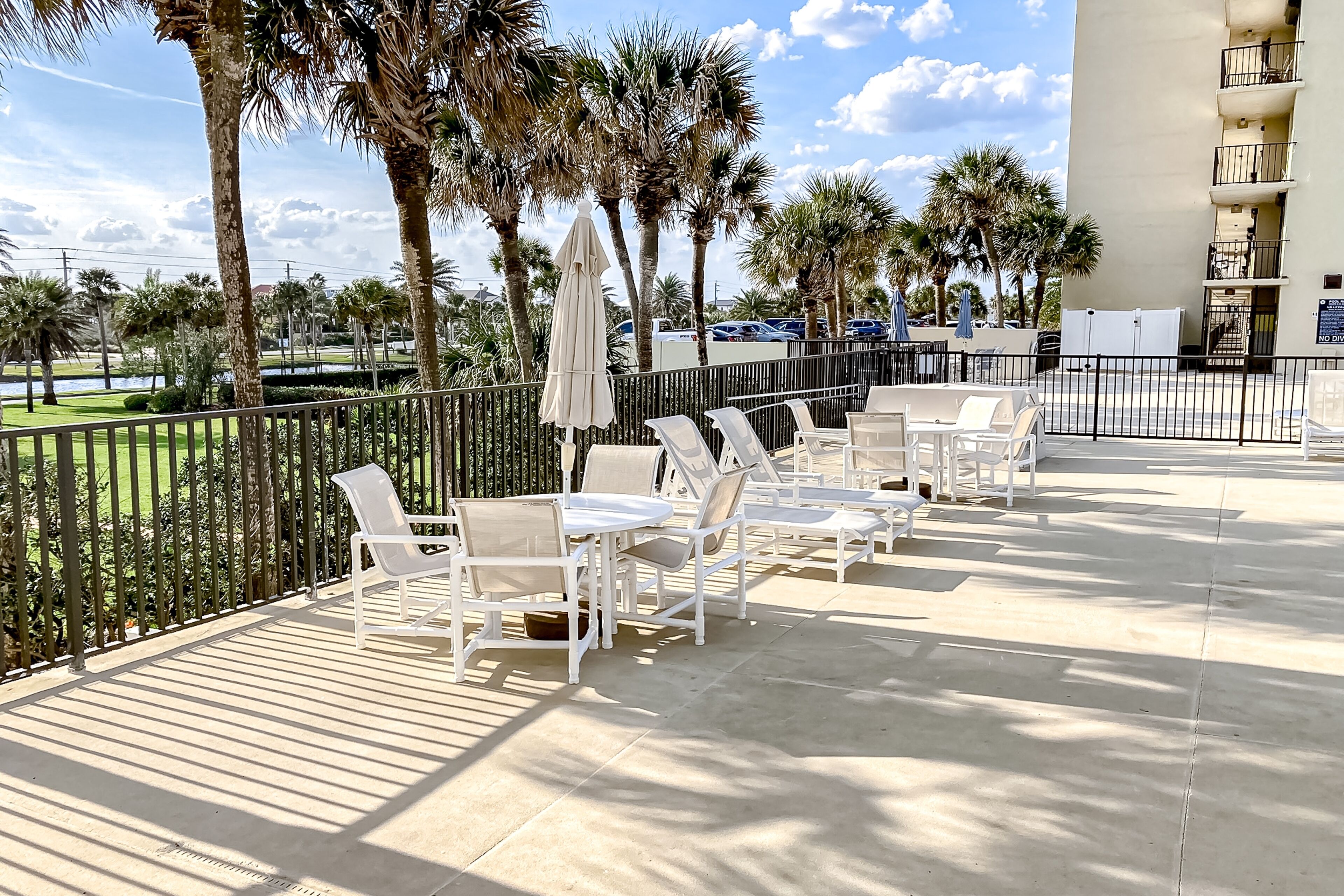 A patio area with white lounge chairs and tables, palm trees, and a view of a parking lot.