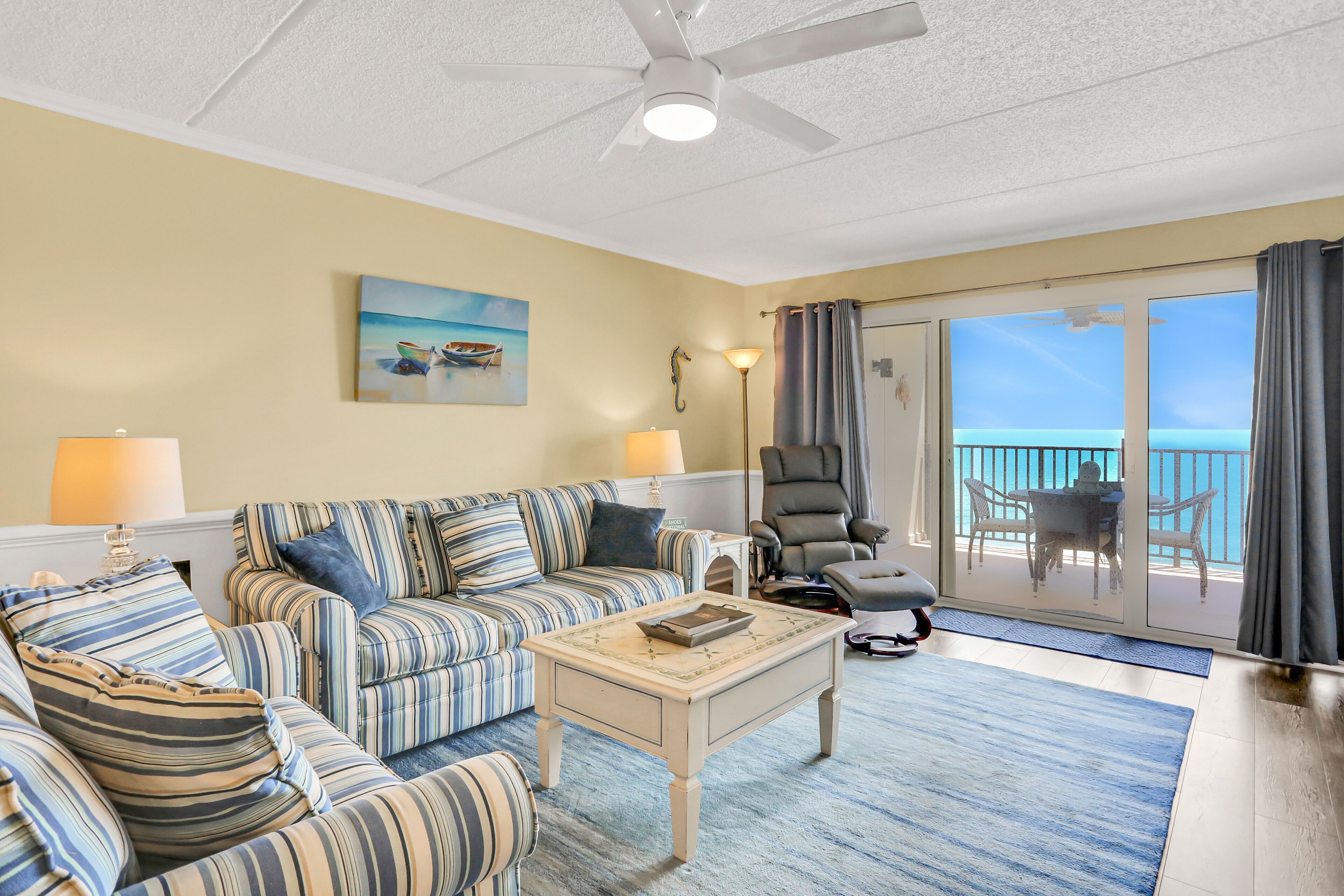 A living room with striped furniture, a ceiling fan, and a view of the ocean through large sliding doors.