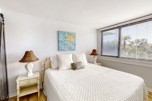 A bedroom with a large bed, two bedside tables, a wall art piece, and a window with blinds. - Spacious Condo With Beach Access, Balcony, AC & Outdoor Pool (Fernandina Beach)