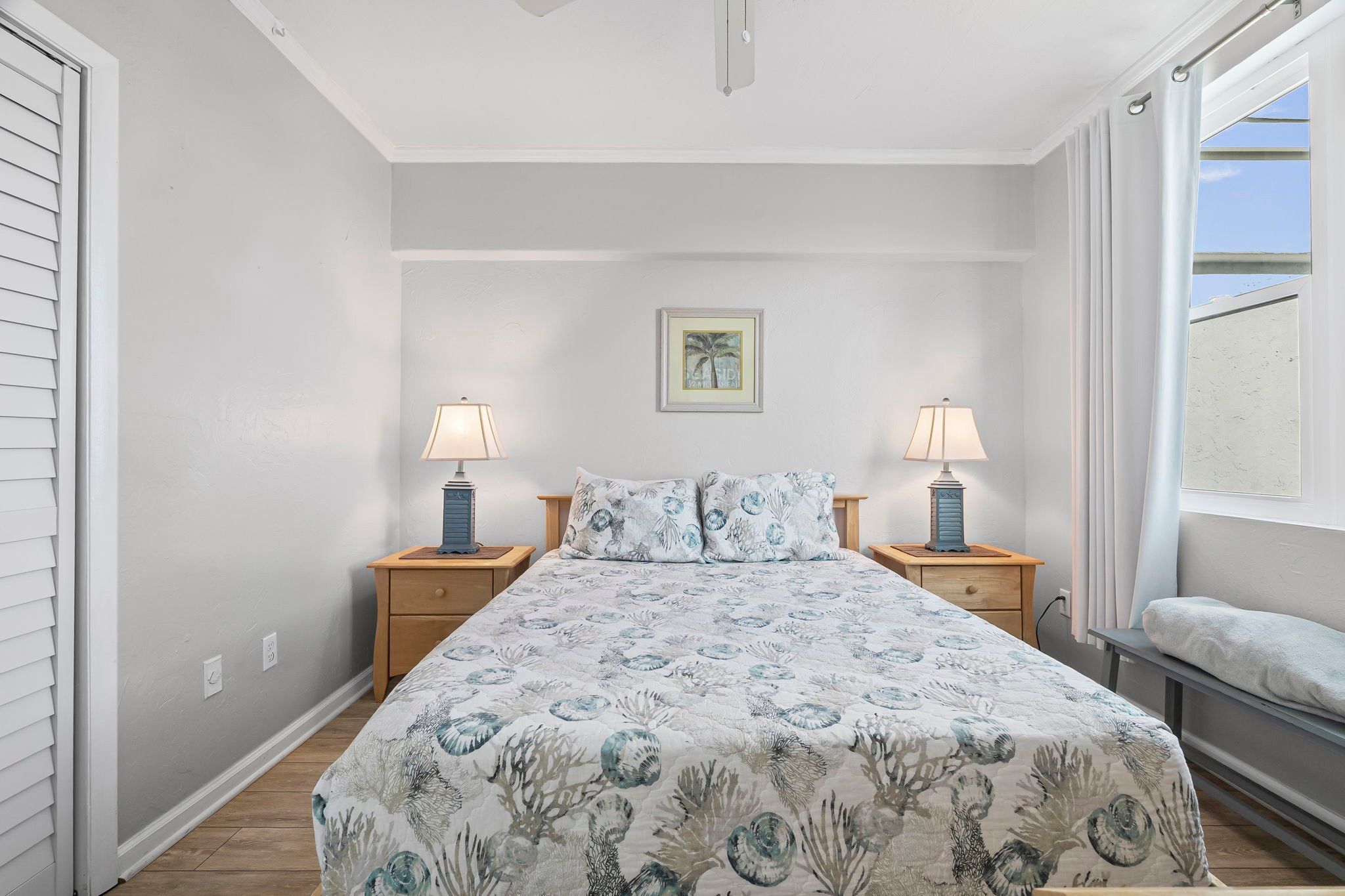 Une chambre à coucher avec un lit, deux tables de chevet avec des lampes, une fenêtre avec des rideaux et un tableau encadré au mur.
