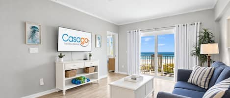 A modern living room with a flat-screen TV, a white coffee table, a blue sofa, and a view of the beach through large windows.