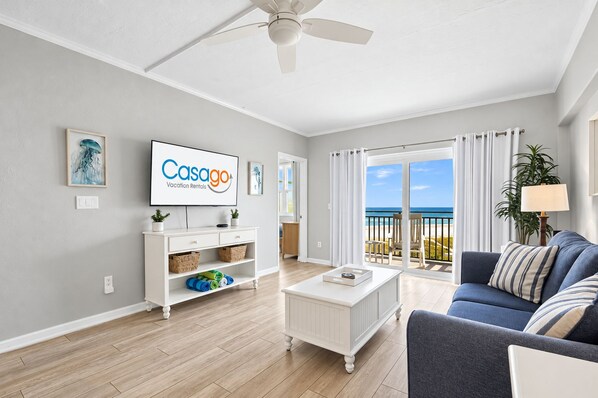 A modern living room with a flat-screen TV, a white coffee table, a blue sofa, and a view of the beach through large windows. - Polished Oceanfront Condo With Pool Access, Private Balcony & Washer/dryer (Jacksonville Beach)
