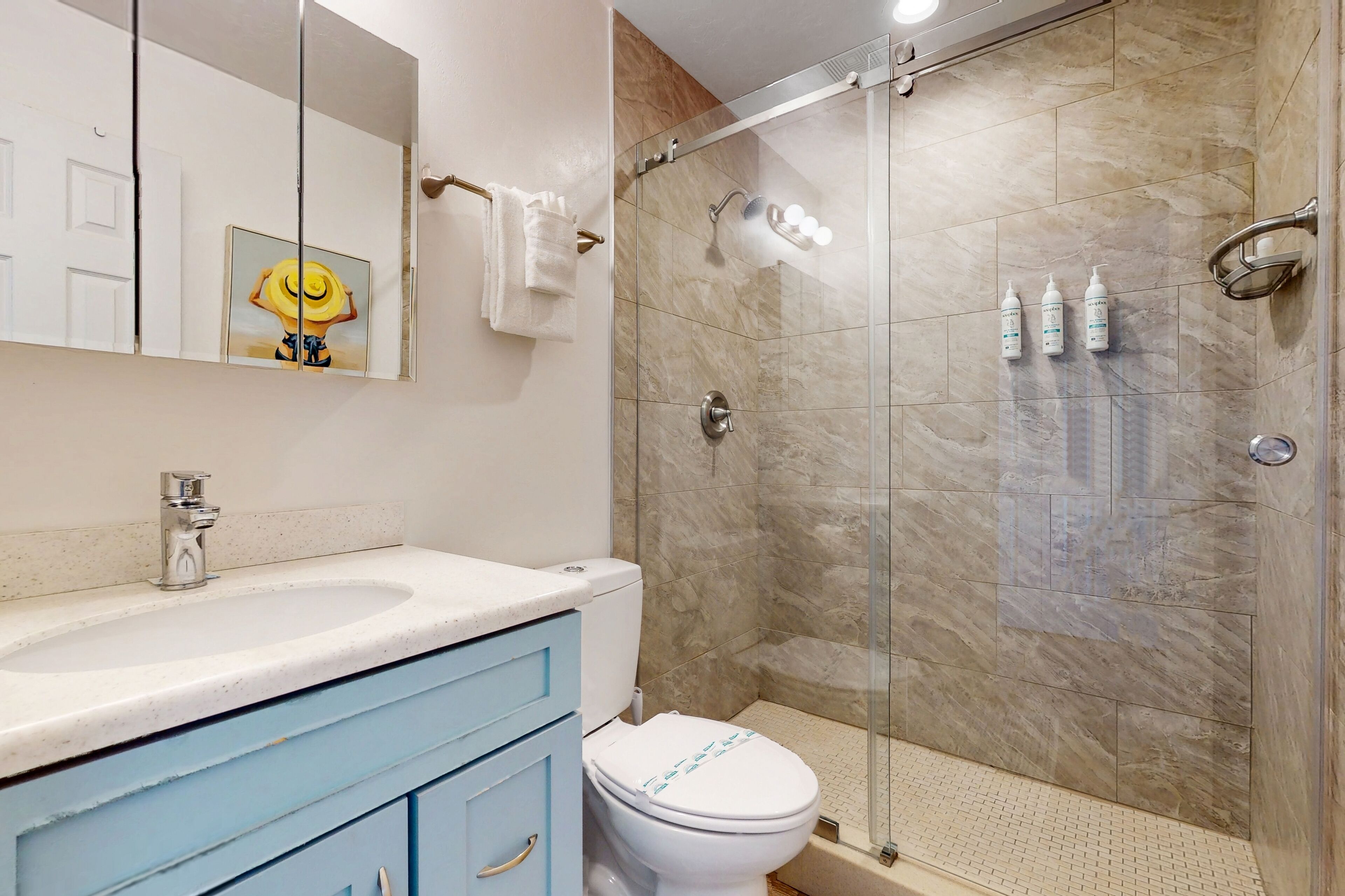 A bathroom with a glass-enclosed shower, a white toilet, a sink with a marble countertop, and a mirror with a painting.