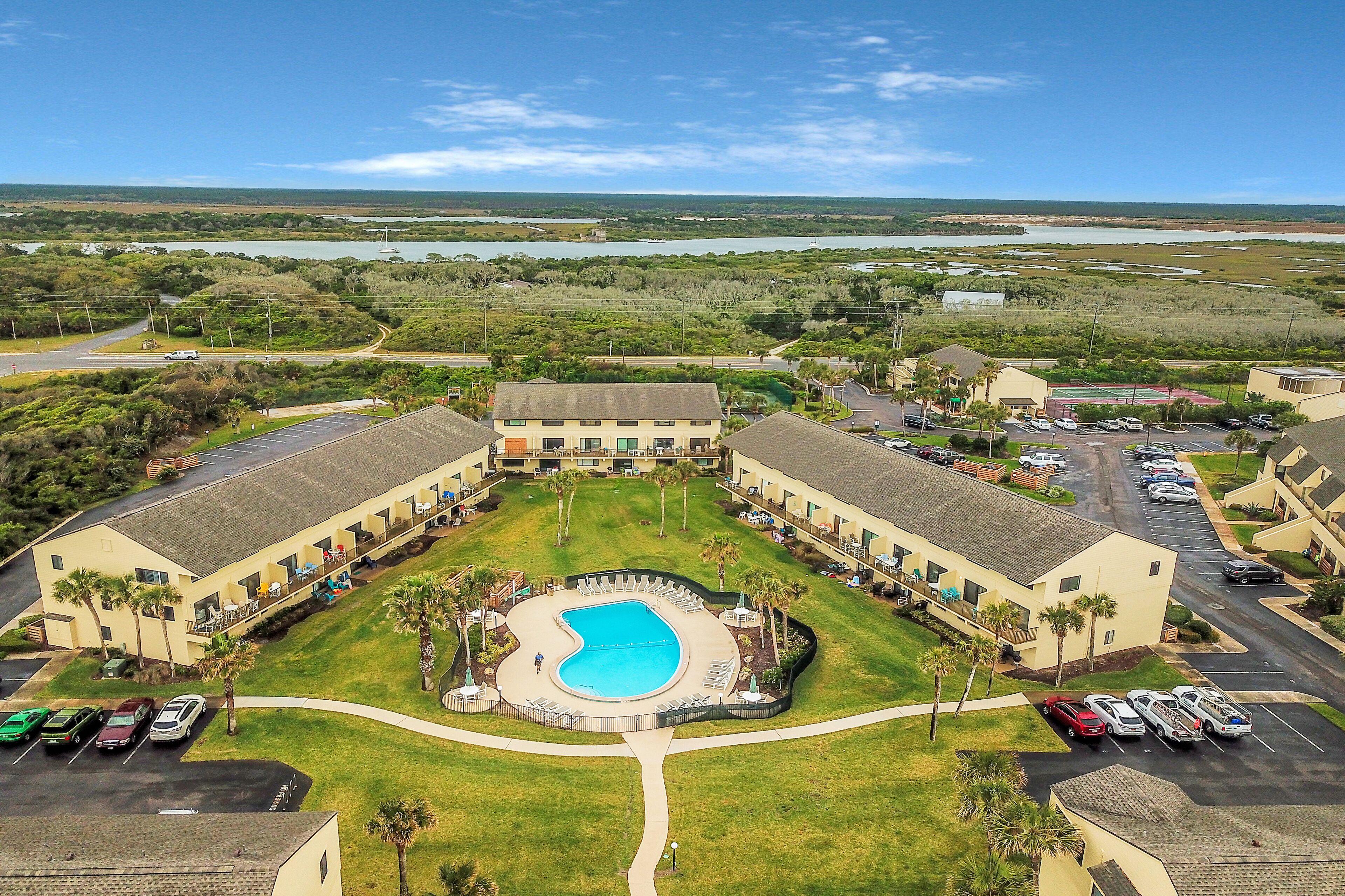 A hotel with a central swimming pool, multiple parking lots, and a view of a lake and forested area.