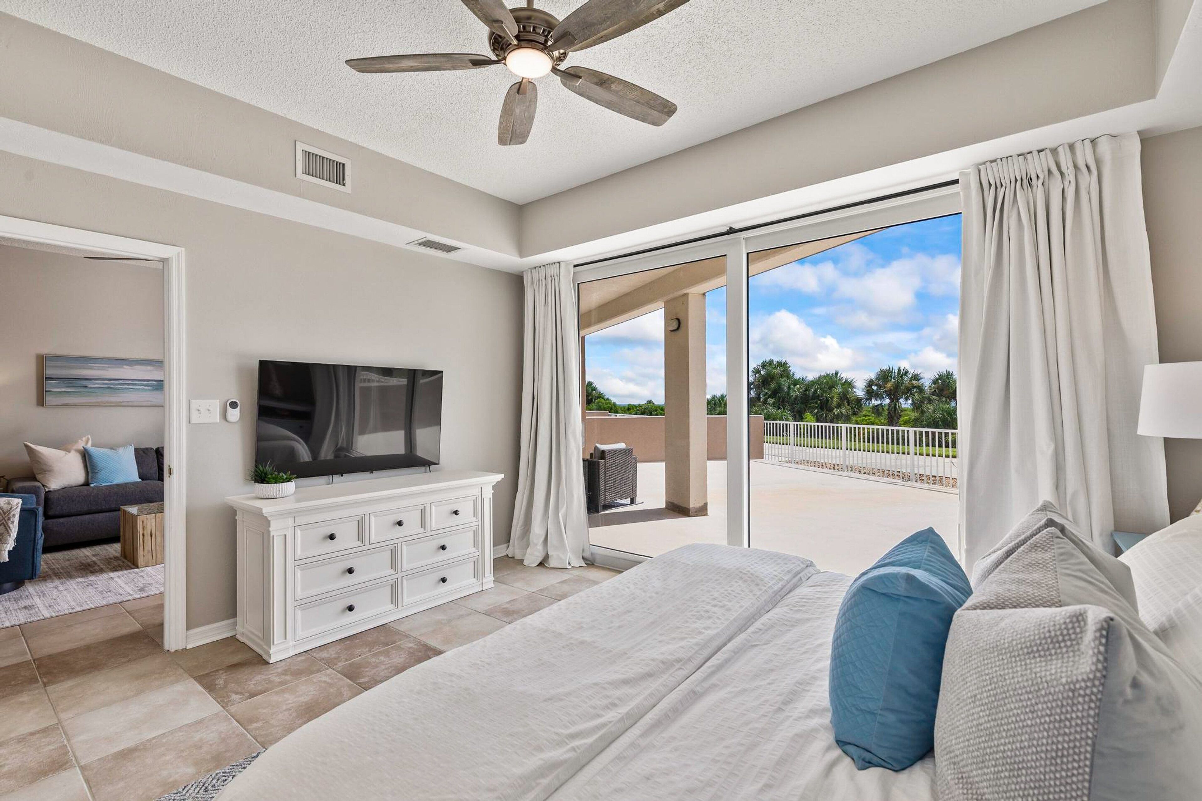 A bedroom with a large bed, a ceiling fan, a TV, and a balcony with curtains.