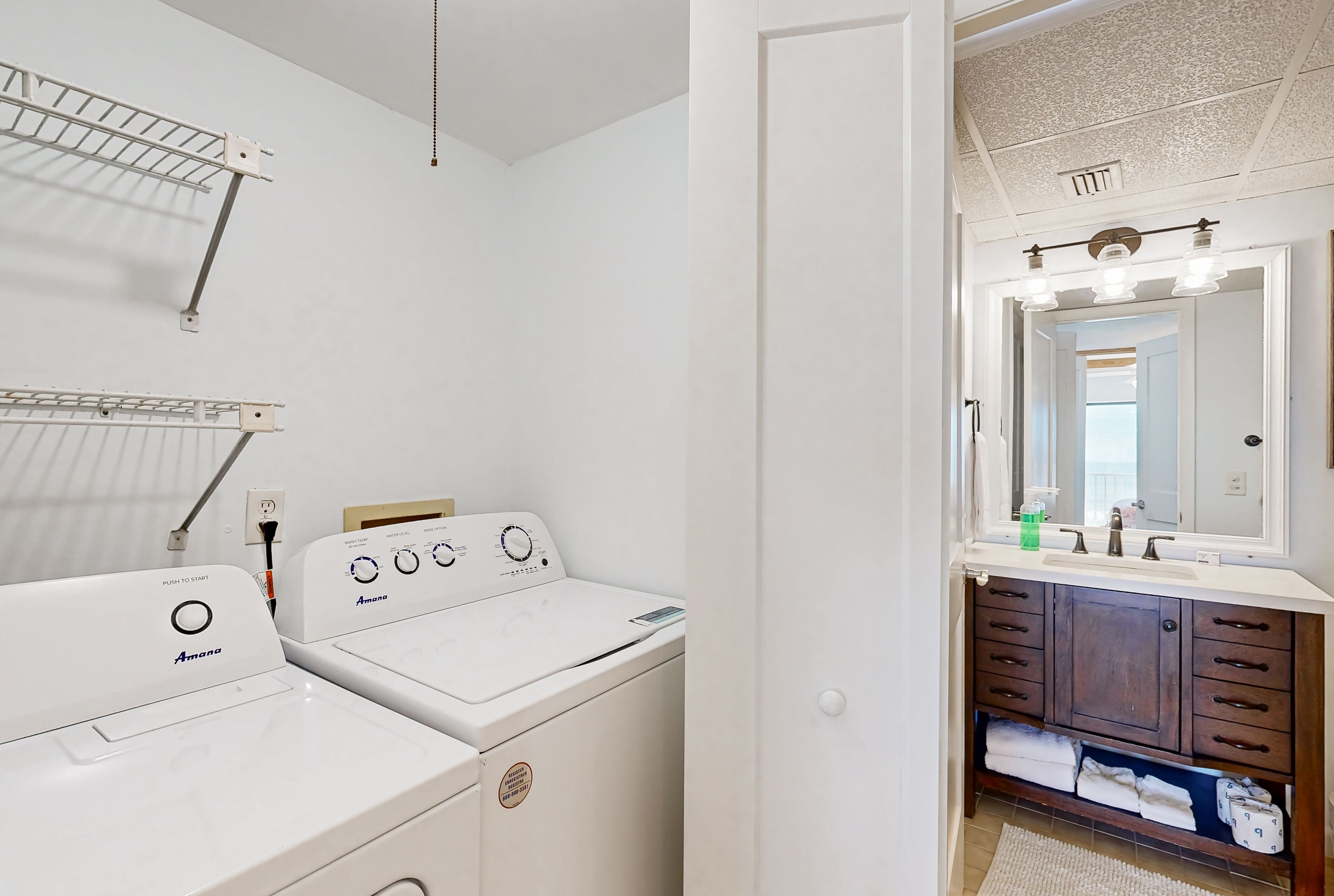 A laundry area with two white Amana washers and dryers, a drying rack, and a bathroom with a vanity, mirror, and towel rack.