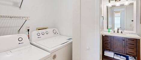 A laundry area with two white Amana washers and dryers, a drying rack, and a bathroom with a vanity, mirror, and towel rack.