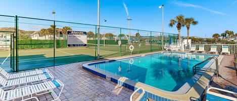 A swimming pool with lounge chairs, a fenced tennis court, and palm trees.