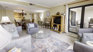 A spacious living room with a sofa, armchairs, a coffee table, and a television. There is a dining area in the background with a table and chairs. - Oceanfront Condo With Pool & Tennis - Balcony, AC, Wifi & Washer/dryer (Fernandina Beach)