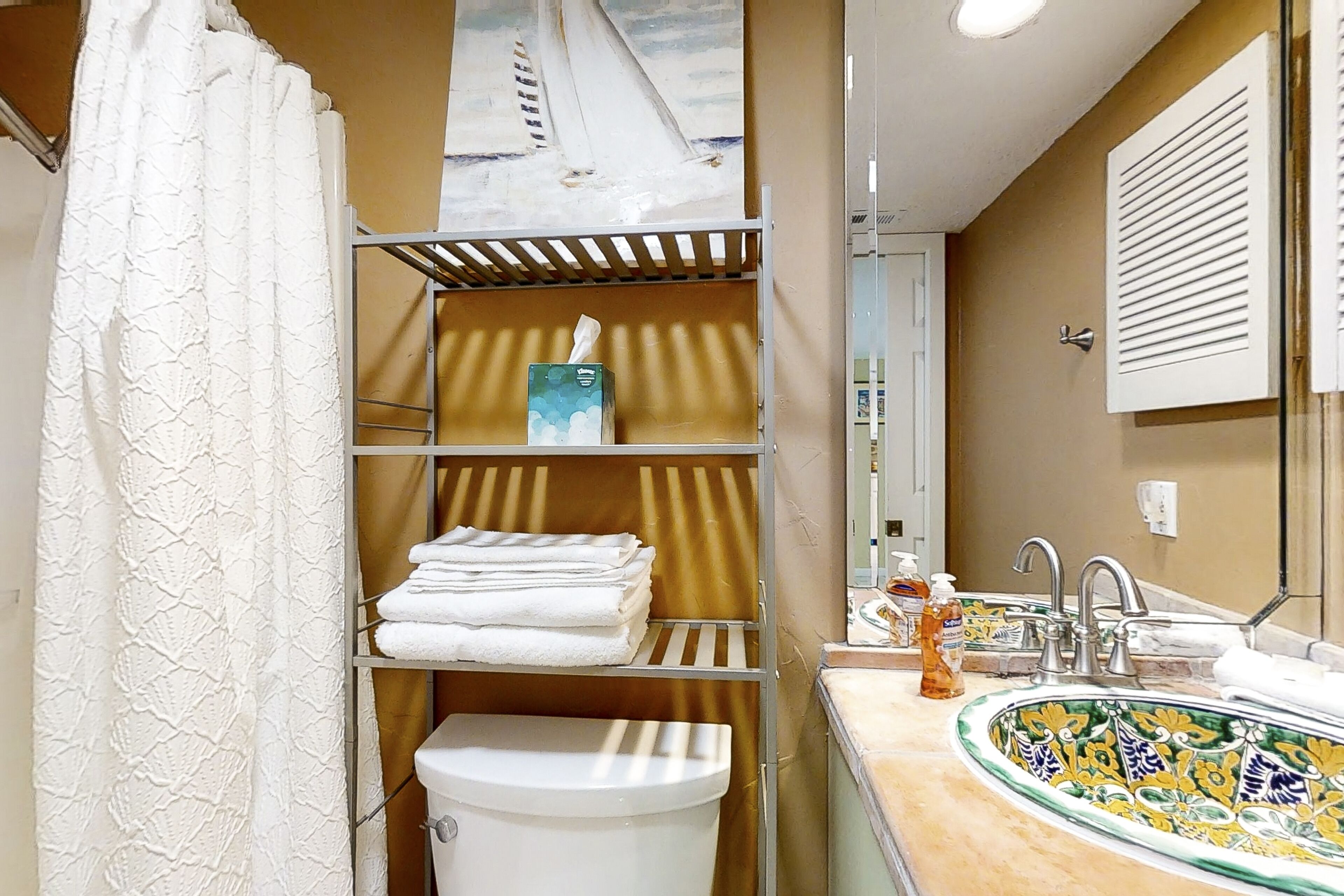 Een badkamer met toilet, planken met opgevouwen handdoeken, een wastafel met decoratieve tegeltjesbak en een douchegordijn.