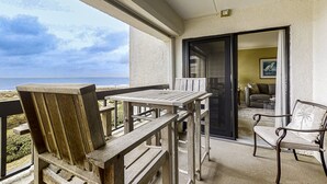 A balcony with a wooden table and chairs, a view of the ocean, and a doorway leading to an interior room with a sofa and a painting. - Oceanfront Condo With Pool & Tennis - Balcony, AC, Wifi & Washer/dryer (Fernandina Beach)