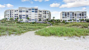 A beachfront hotel with multiple floors, a sandy beach in the foreground, and palm trees. - Oceanfront Condo With Pool & Tennis - Balcony, AC, Wifi & Washer/dryer (Fernandina Beach)
