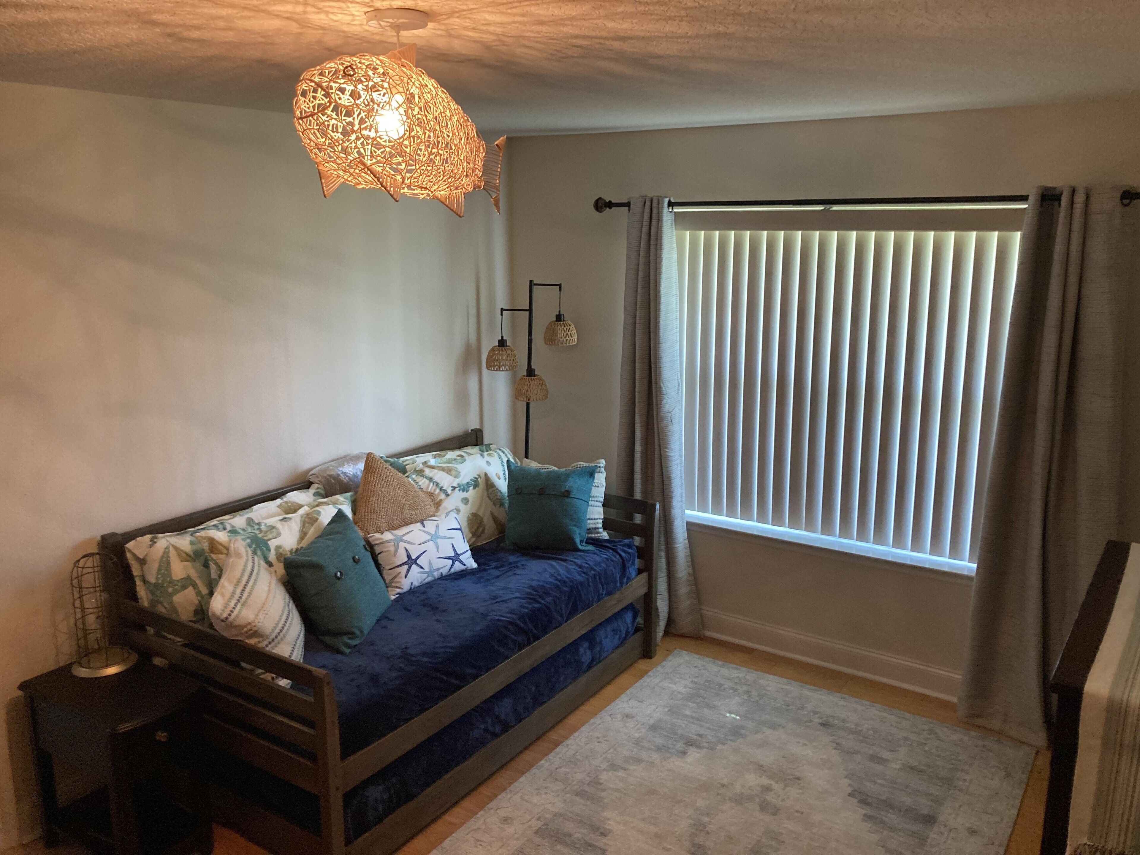 A cozy room with a bed, a hanging light fixture, a window with blinds, and a small table.
