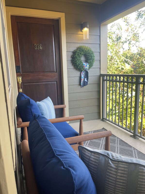 A wooden door with a number, a porch with a bench and cushions, a wreath, and a railing with greenery in the background.