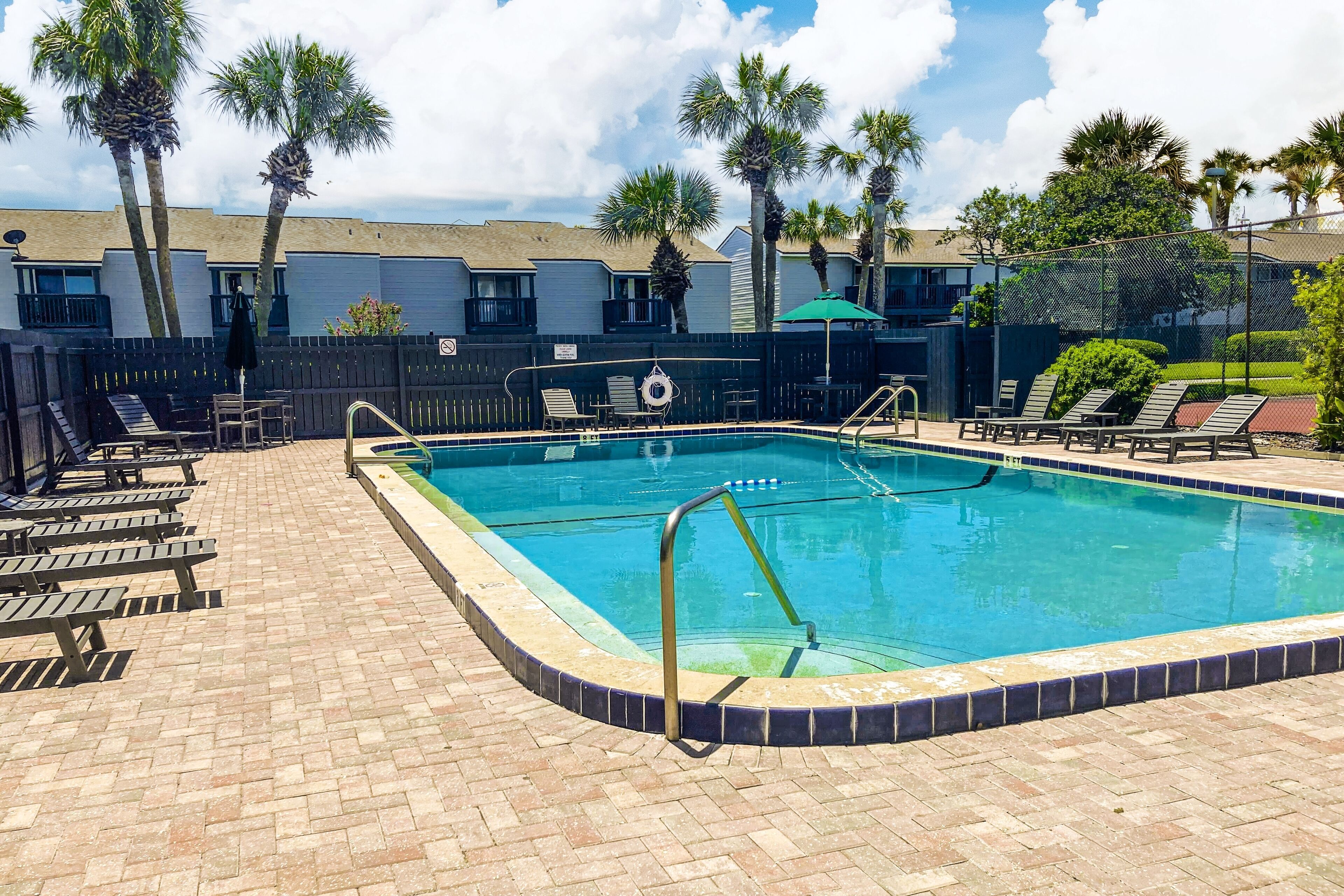 Une piscine rectangulaire dont le bord est recouvert de carreaux, entourée d’une terrasse en briques. On y trouve des chaises longues et un poste de surveillance pour les sauveteurs, près de la piscine.