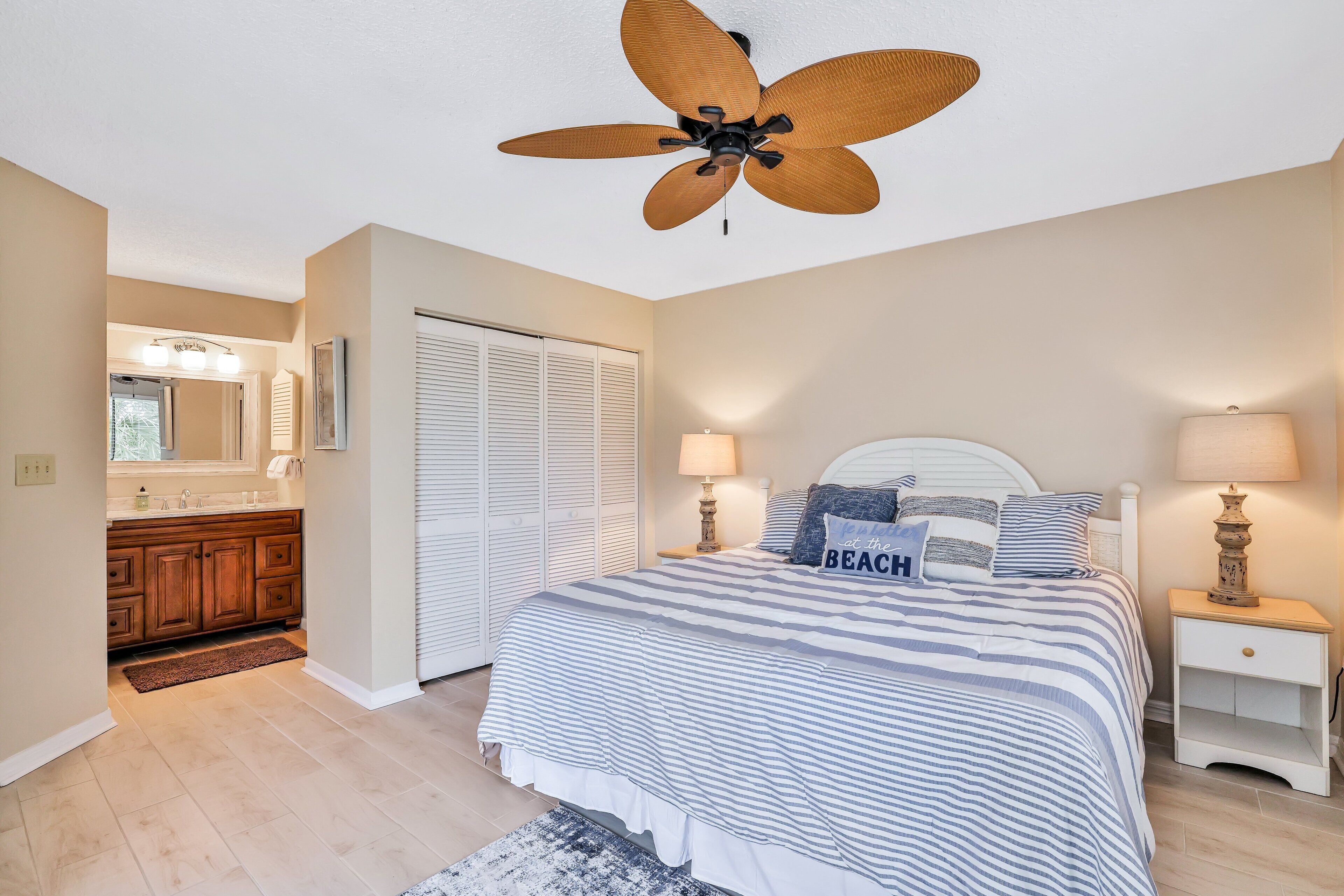 Une chambre à coucher comprenant un lit, des tables de chevet, un ventilateur de plafond et une salle de bain avec un meuble-lavabo, ainsi qu’une porte.