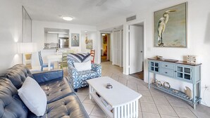 A living room with a black leather sofa, a patterned armchair, and a white coffee table. There is a blue console table with decorative items and a painting of a bird on the wall. - Sunny Ground-floor Condo W/private Patio, W/d, On-site Pools, Tennis Court, BBQ (St. Augustine)