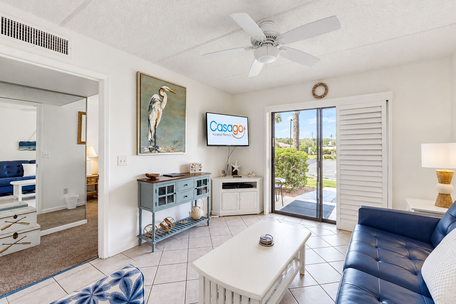 A modern living room with a blue sofa, a white coffee table, a painting of a bird, and a TV displaying 'Casago Vacation Rental'.