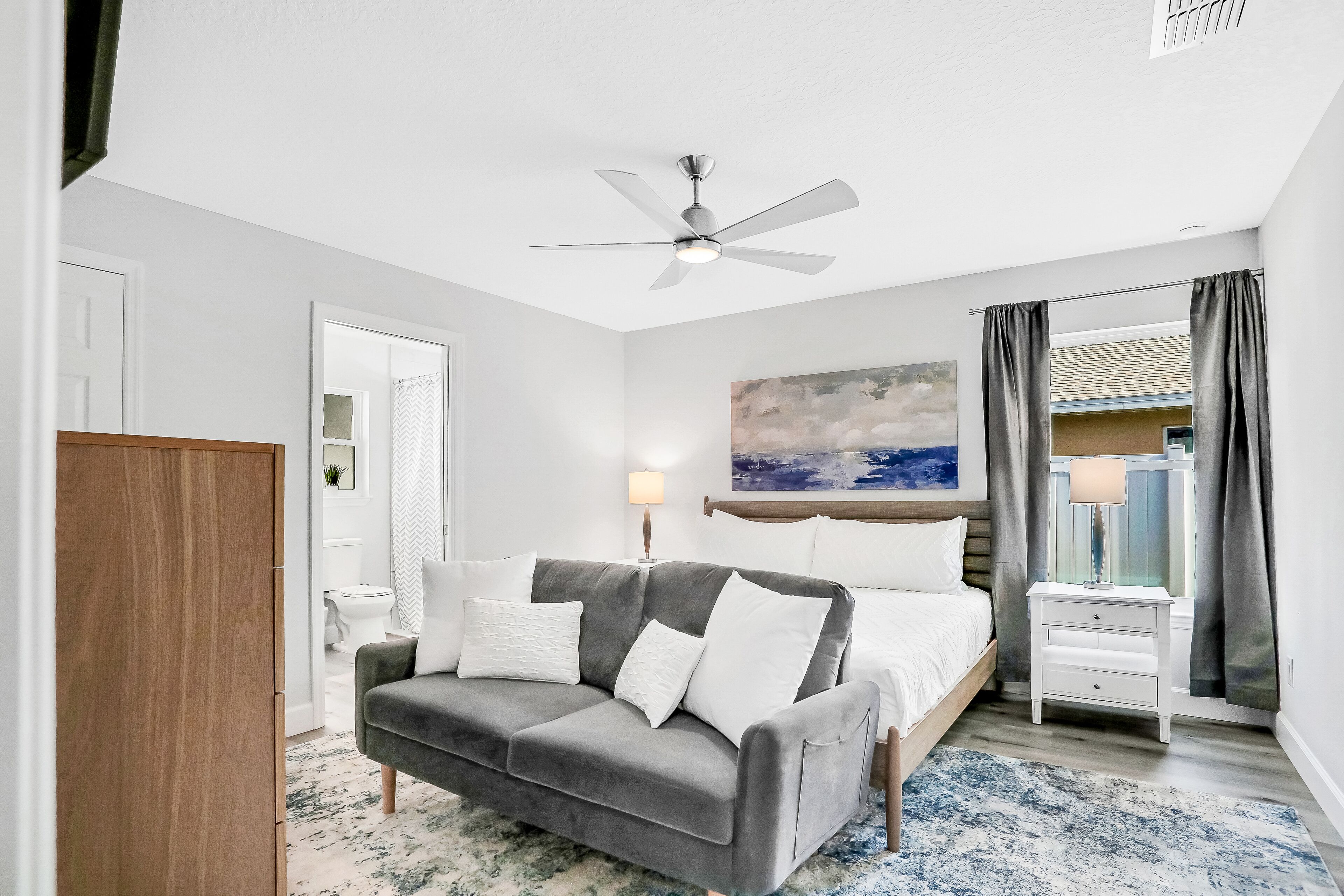 A bedroom with a grey sofa, a bed, a nightstand, a ceiling fan, and a painting on the wall.