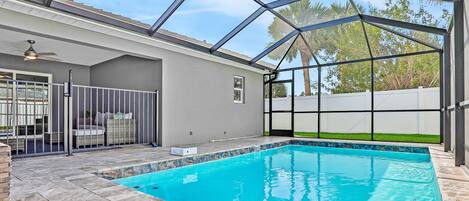A covered swimming pool with a glass ceiling, a patio area with seating, and a view of palm trees.