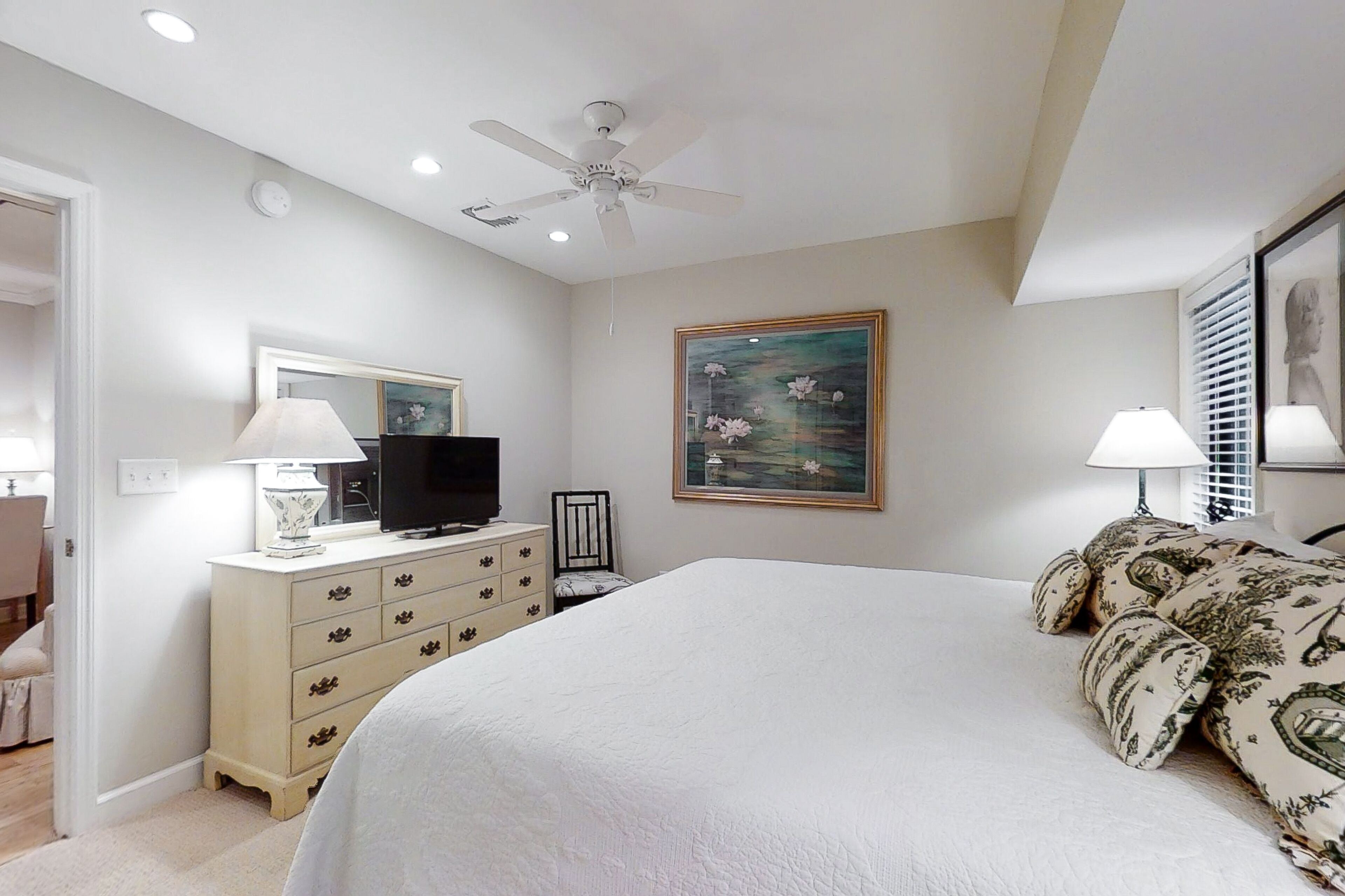 A bedroom with a bed, dresser, TV, ceiling fan, and artwork.