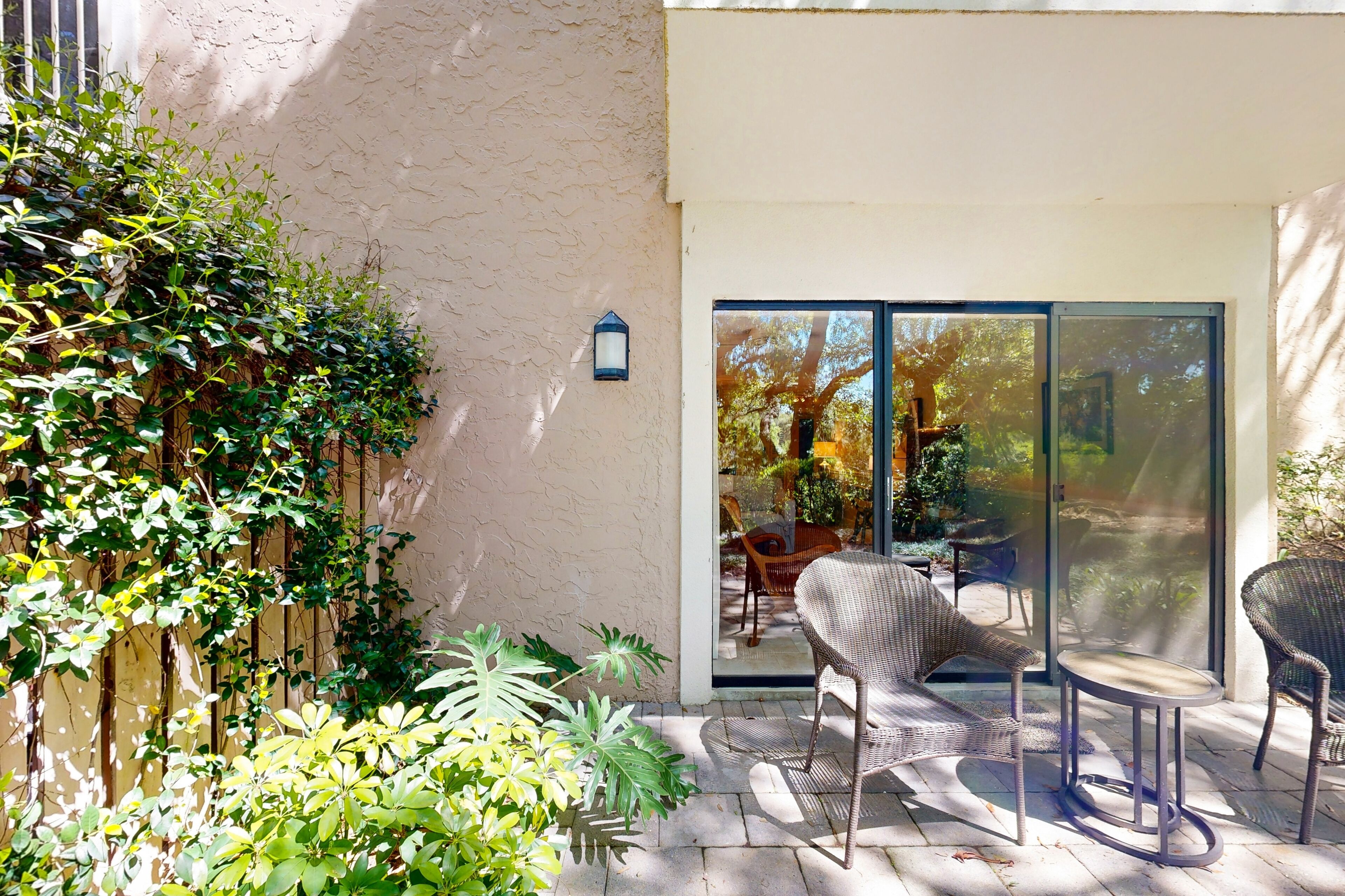 Un patio con mobiliario de mimbre, una puerta de vidrio que da a un jardín y un farol fijado a la pared.