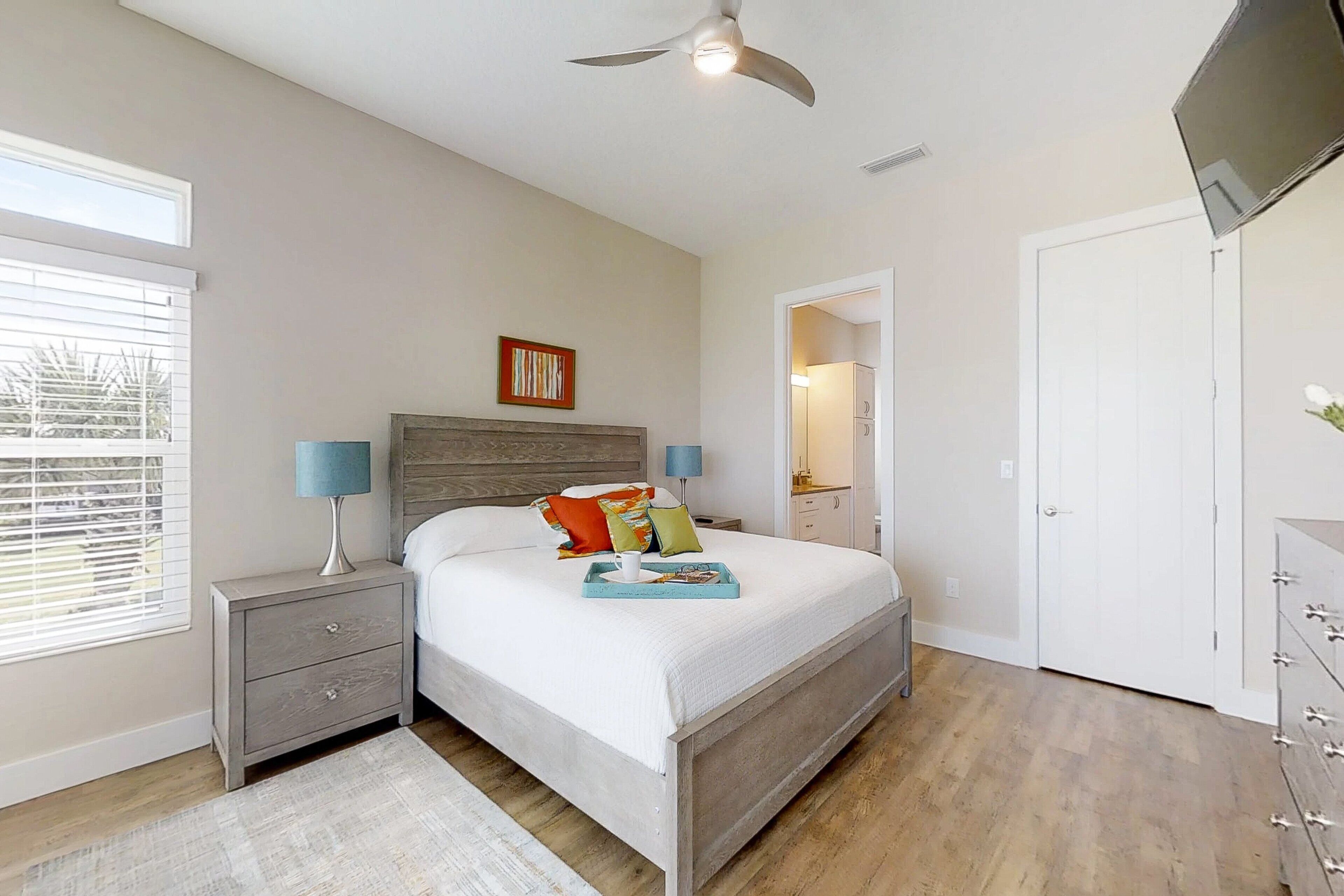 A bedroom with a bed, nightstands, a ceiling fan, a window with blinds, and a TV mounted on the wall.