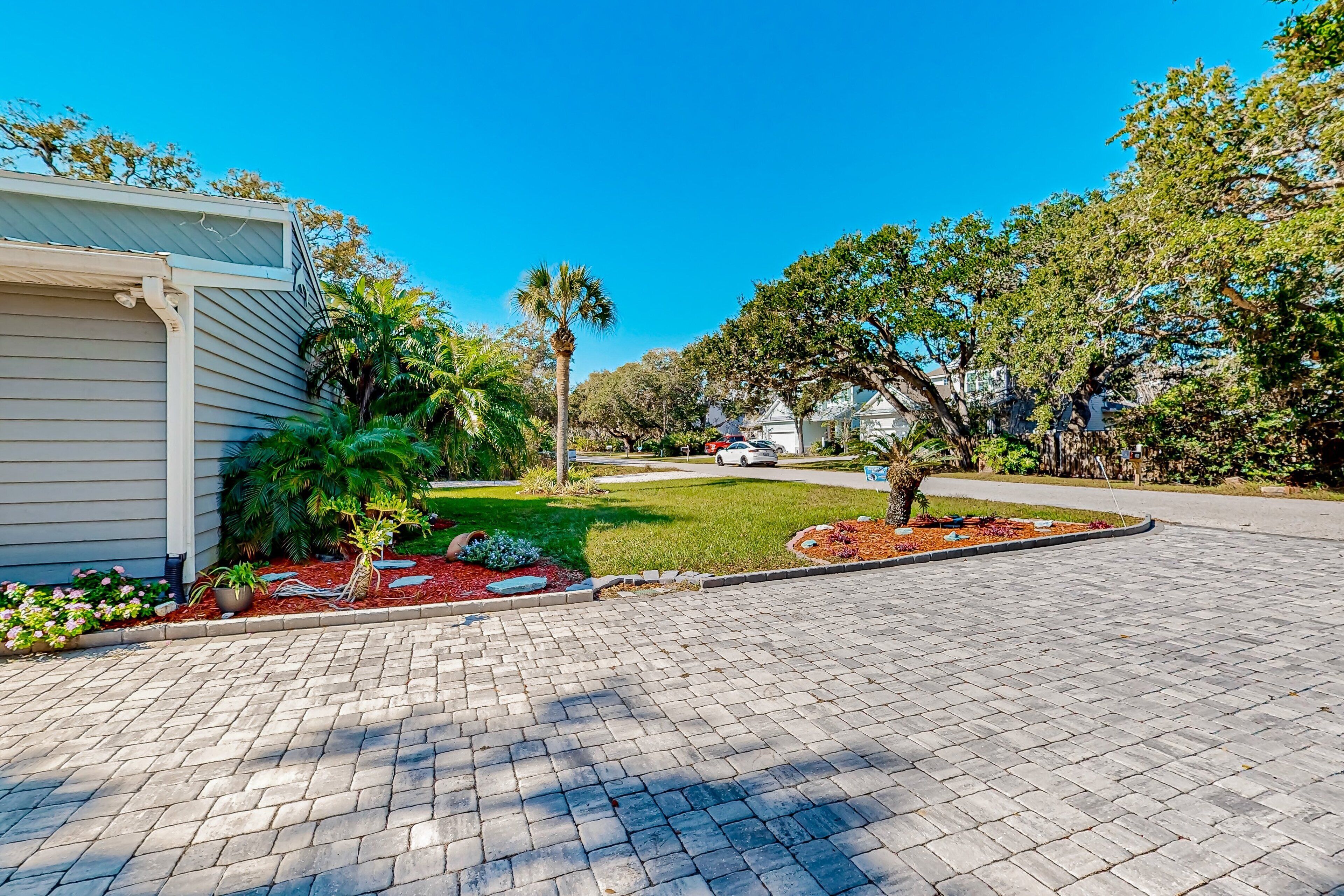 A residential driveway with a paved path, a lawn, and a variety of plants including a palm tree and shrubs.