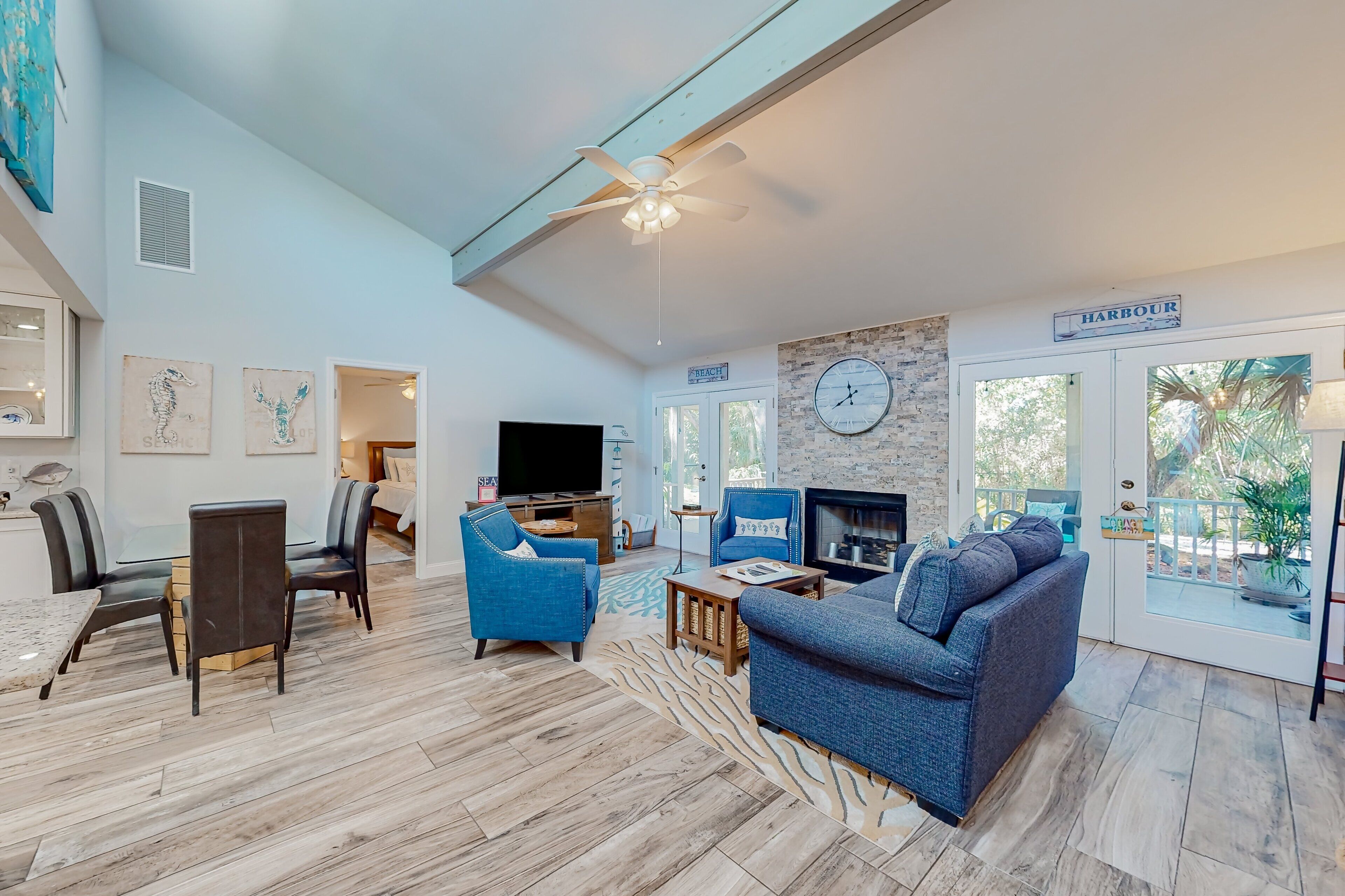 A spacious living room with a large sofa, a television, and a fireplace. There is a dining area with a table and chairs, and a kitchen area with a refrigerator and microwave. The room has a high ceiling with a fan and a view of the outside through large windows.