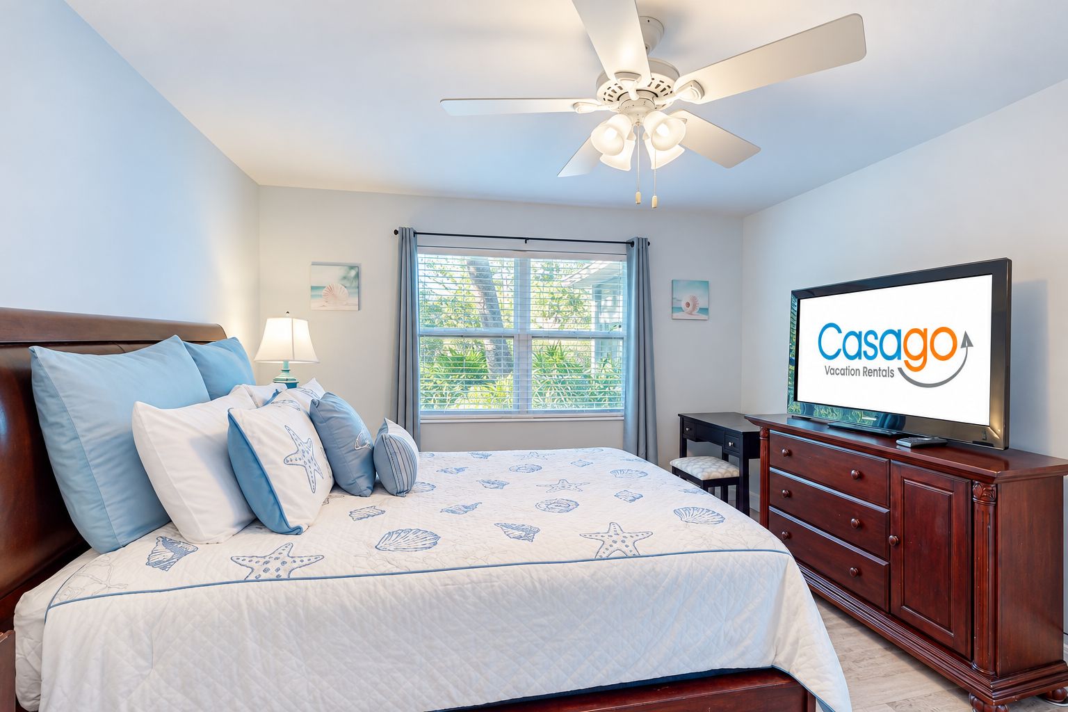 A bedroom with a bed, a ceiling fan, a TV, and a window with a view of greenery.