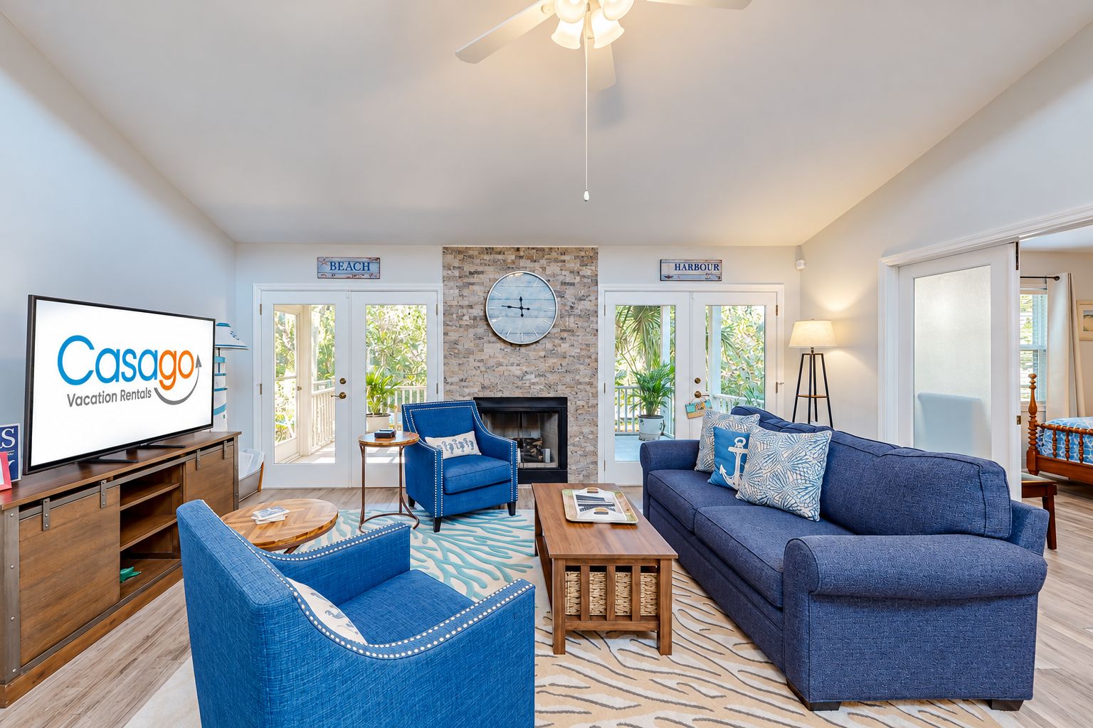 A living room with a television displaying 'Casago Vacation Rentals,' a fireplace, a clock, and a view of the outdoors.