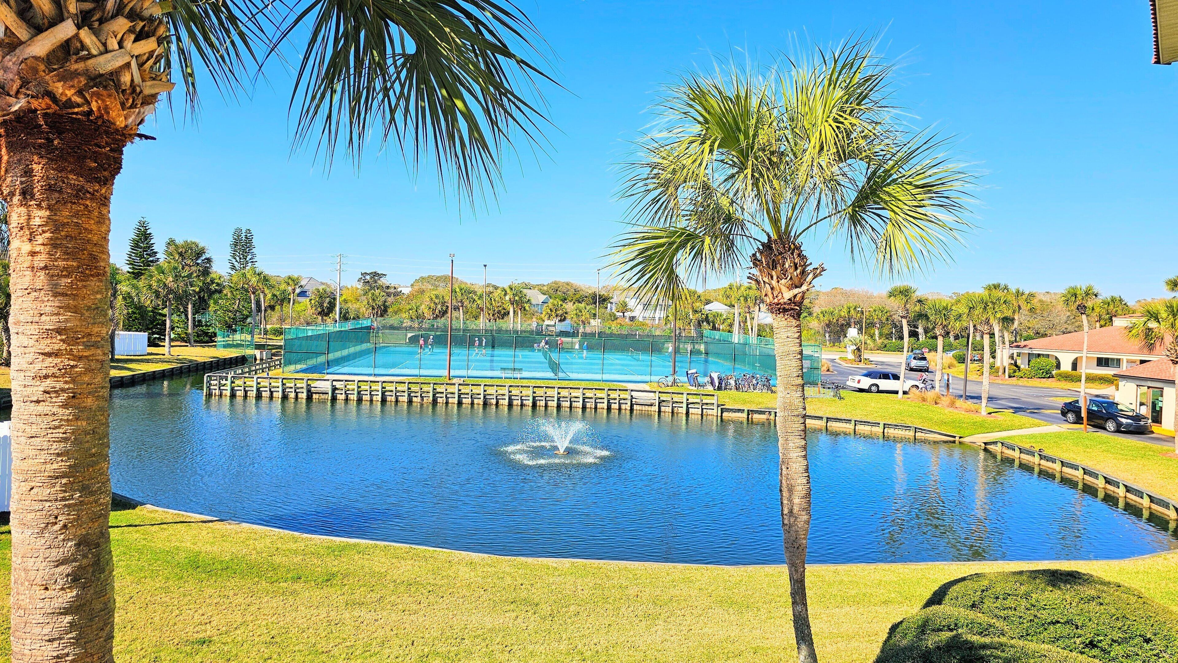 A tennis court adjacent to a calm water body with a fountain, surrounded by palm trees and a clear sky.