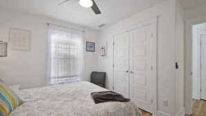 Ein Schlafzimmer mit Bett, Sessel, Deckenventilator, Fenster mit Jalousien und einer Tür.