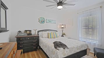 Une chambre à coucher comprenant un lit, une table de chevet, une commode, un ventilateur de plafond, une fenêtre avec des rideaux et un mur orné de deux tableaux encadrés.