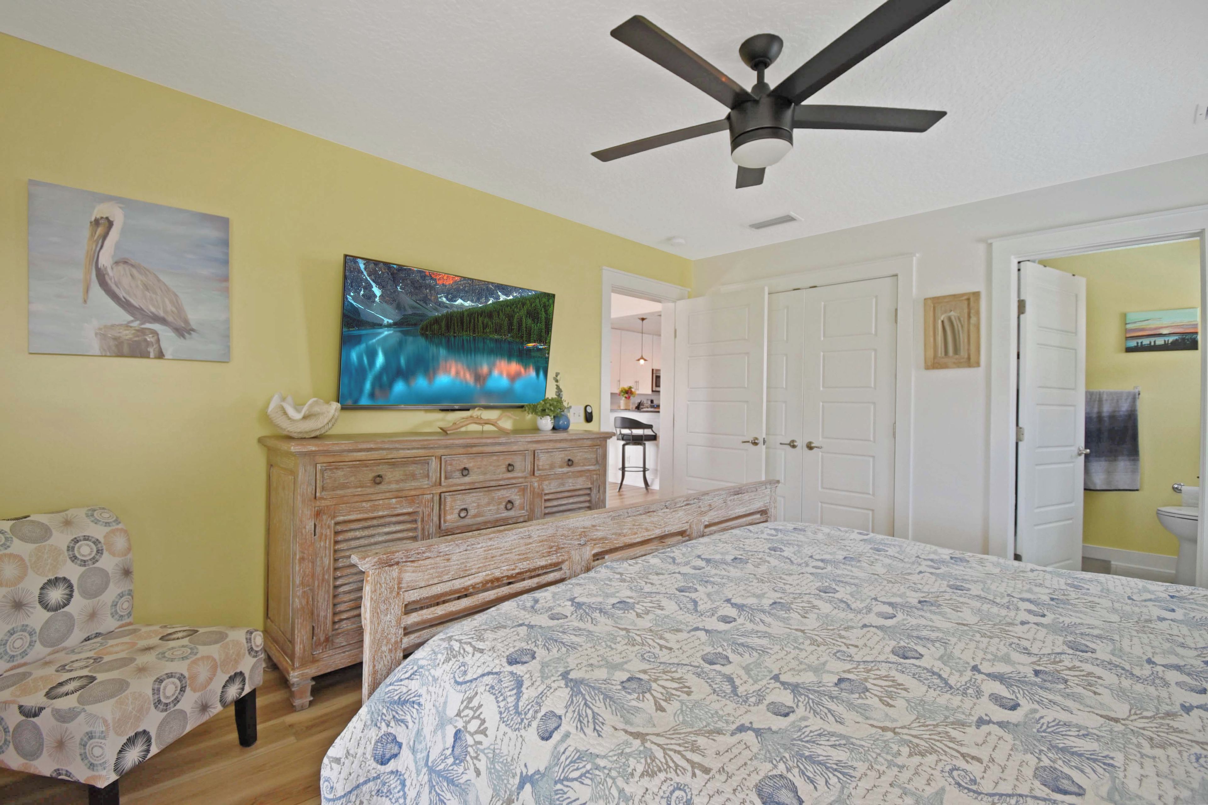 A bedroom with a bed, a dresser, a chair, a ceiling fan, and a painting of a mountain lake.