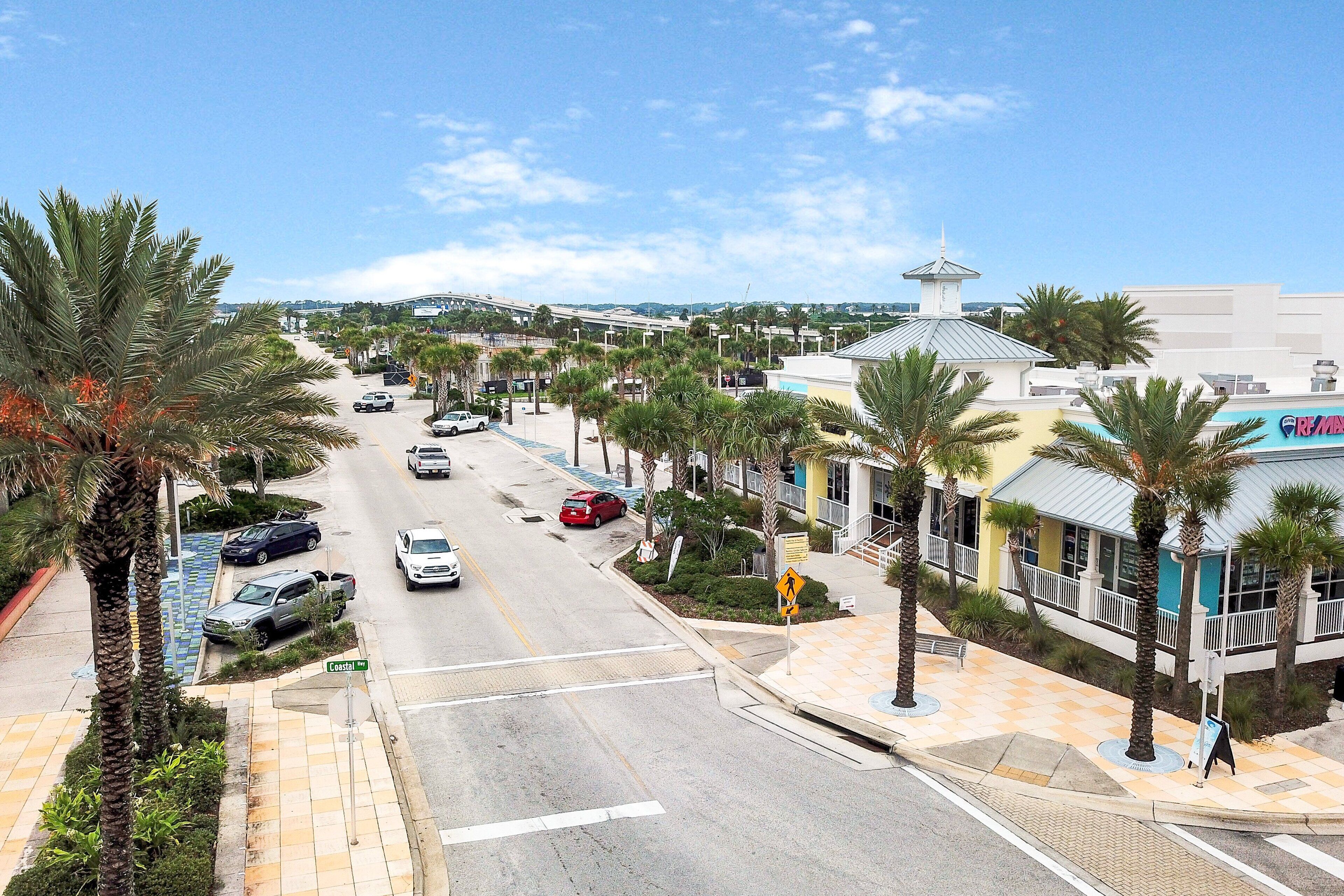 A street view with palm trees, parked cars, and a building with a blue sign.