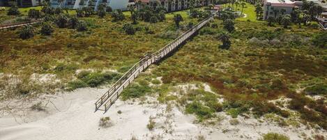 An aerial view of a resort with a long wooden bridge connecting buildings, surrounded by sand dunes and sparse vegetation.