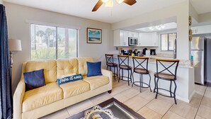 A modern living room with a beige sofa, two bar stools, a ceiling fan, and a kitchen area with white cabinets and stainless steel appliances. - Sun-kissed Beach Condo With Amazing Pool, Tennis & Patio - Steps to the Beach! (St. Augustine)