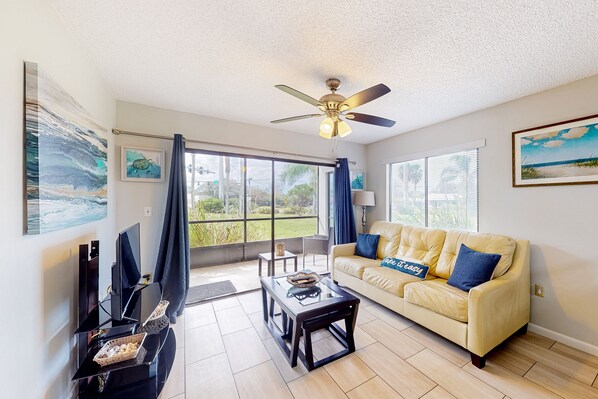 A living room with a sofa, coffee table, TV, and artwork. - Sun-kissed Beach Condo With Amazing Pool, Tennis & Patio - Steps to the Beach! (St. Augustine)