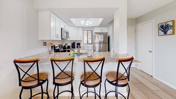 A modern kitchen with white cabinets, a granite countertop, and a central island with bar stools. - Sun-kissed Beach Condo With Amazing Pool, Tennis & Patio - Steps to the Beach! (St. Augustine)