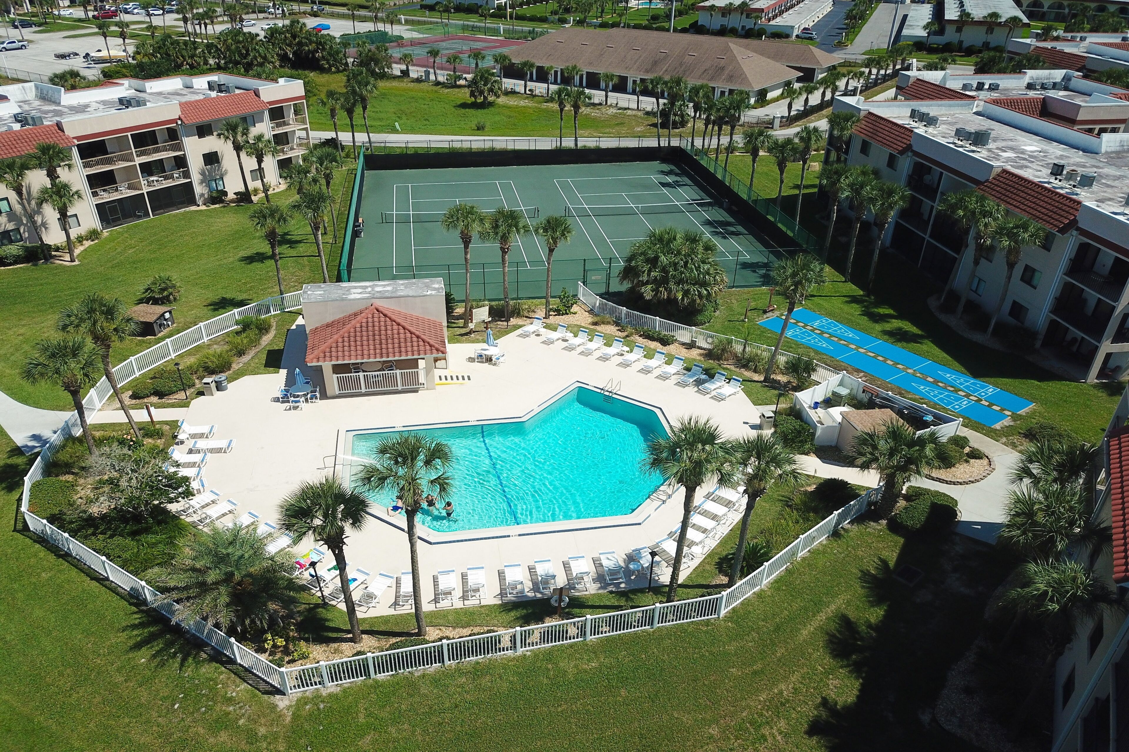 Vista aérea de un hotel con piscina, canchas de tenis y una zona ajardinada con palmeras.