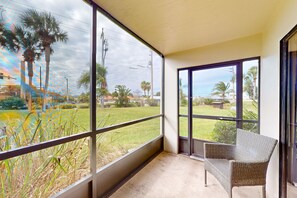 A balcony with a wicker chair, large windows, and a view of palm trees and a grassy area. - Sun-kissed Beach Condo With Amazing Pool, Tennis & Patio - Steps to the Beach! (St. Augustine)