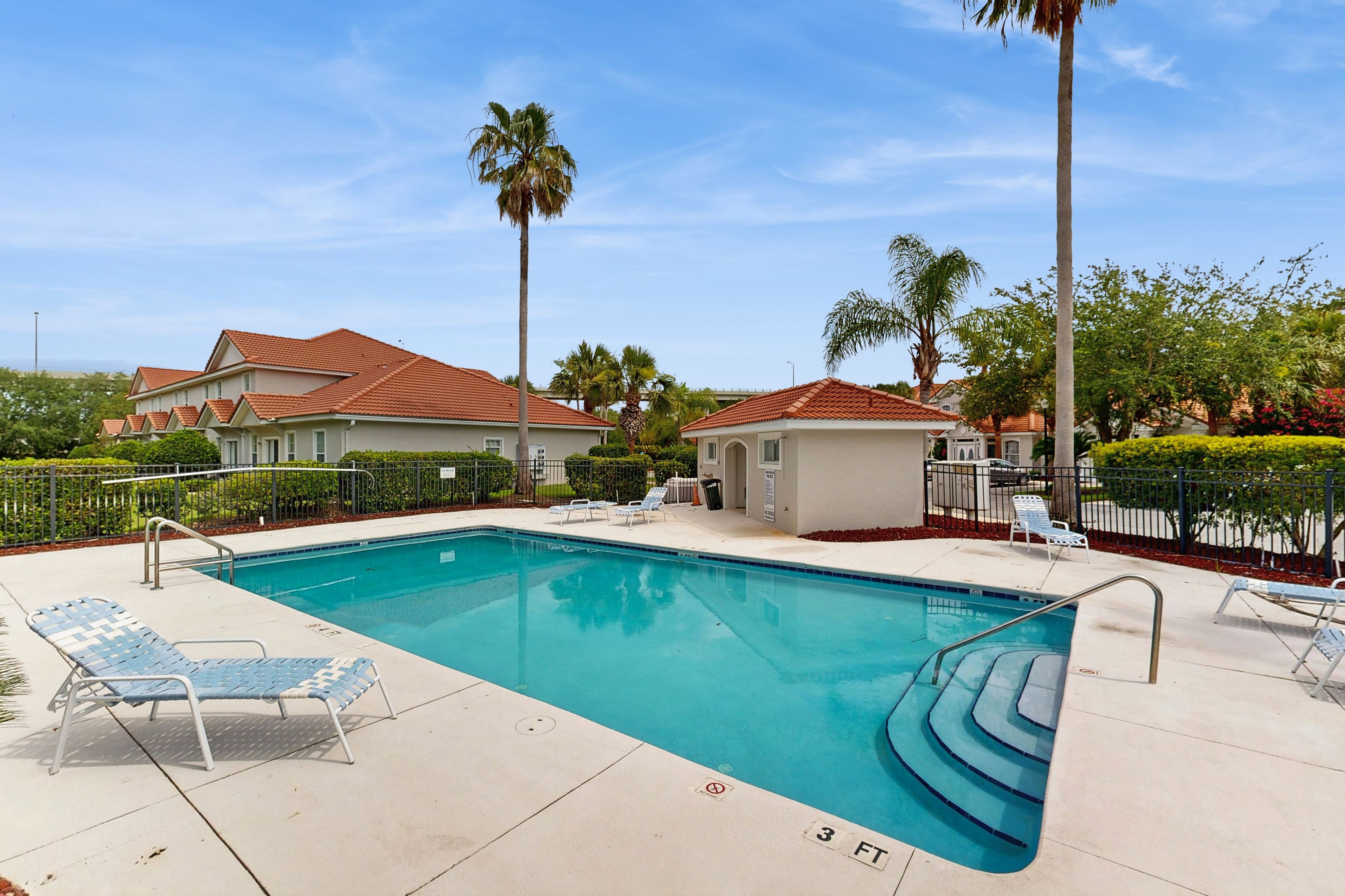 A swimming pool with lounge chairs, palm trees, and a building with red roofs.