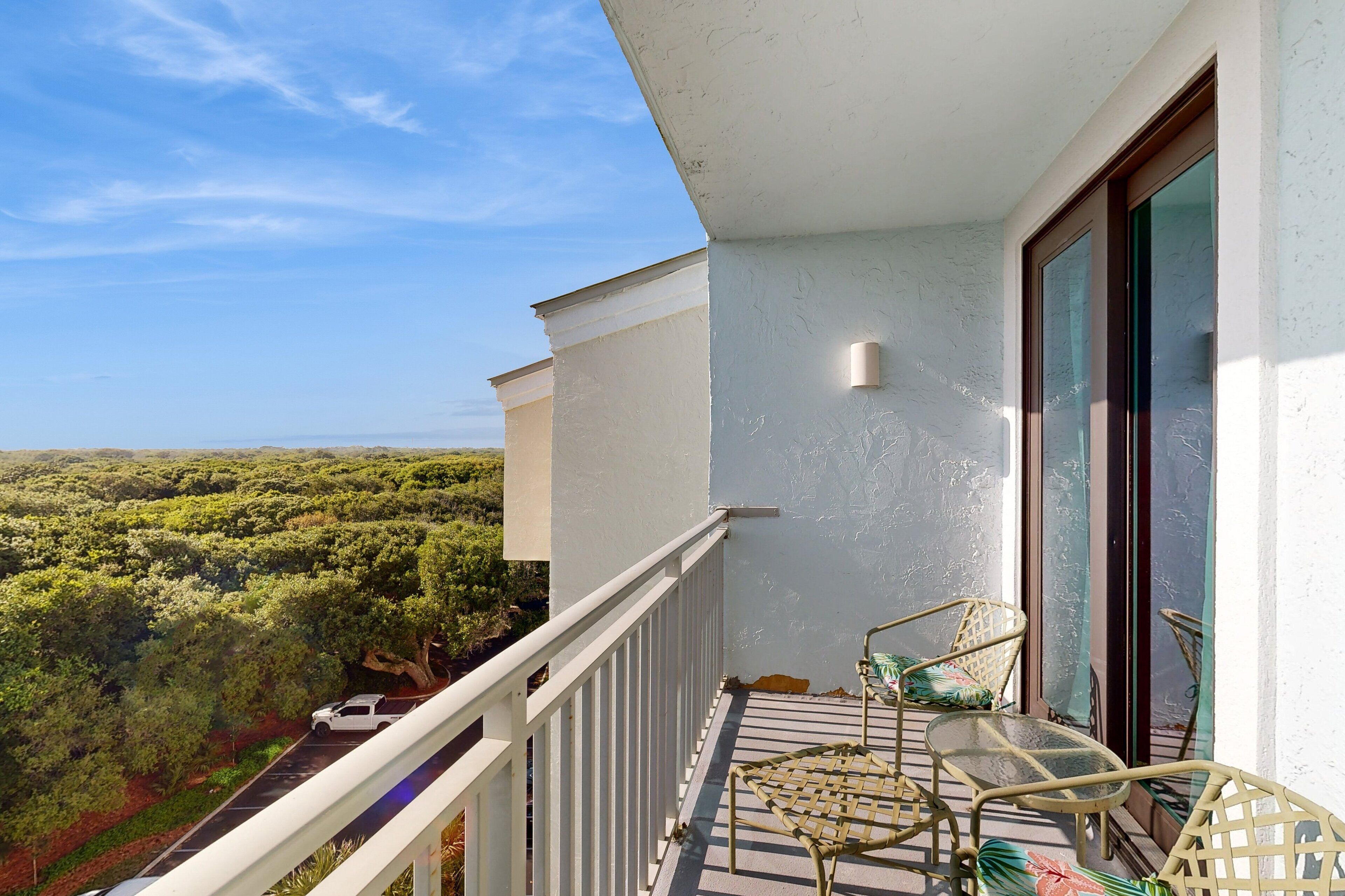 A balcony with a view of a forested landscape, a white railing, and two wicker chairs.