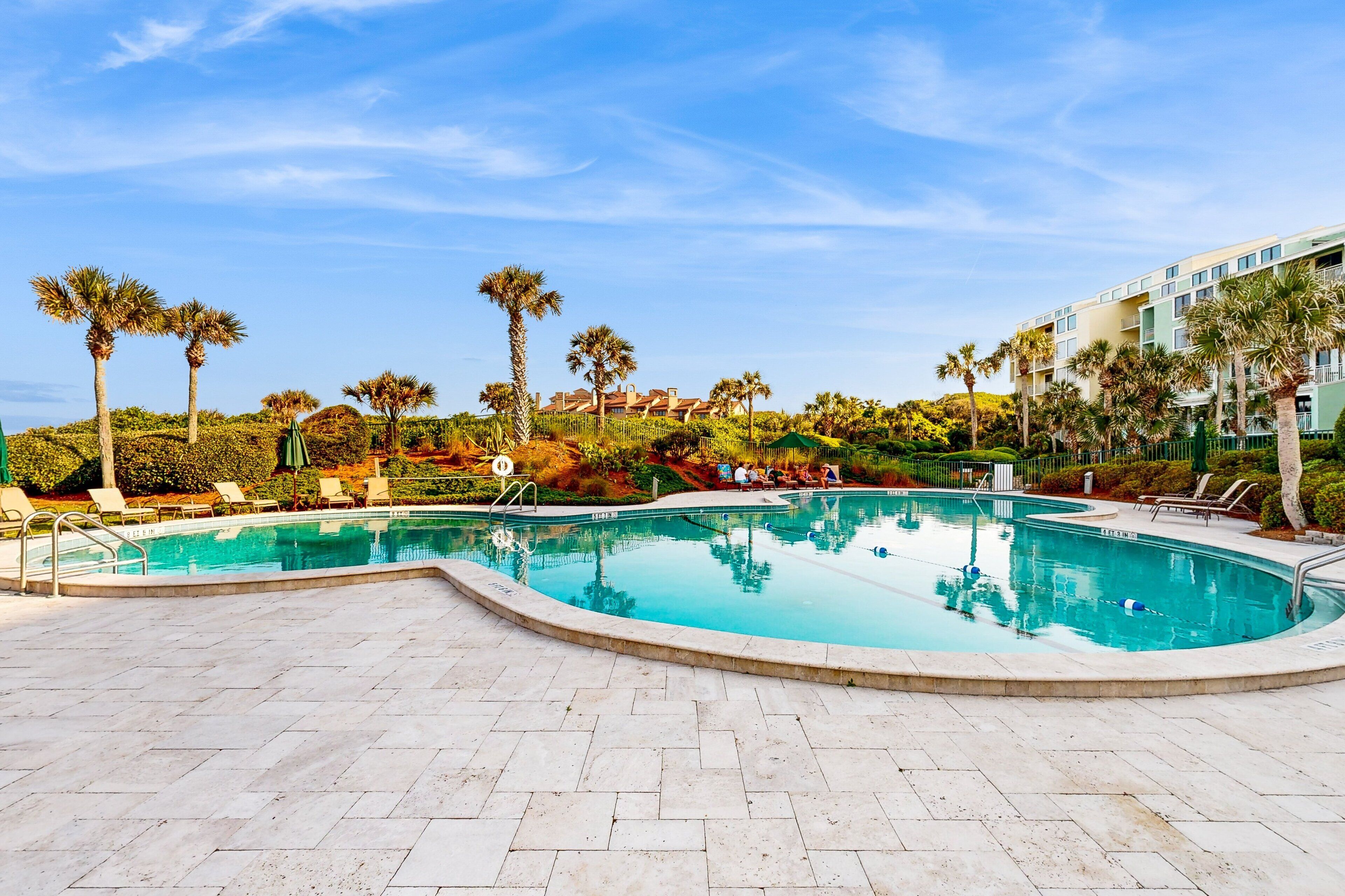 A swimming pool with lounge chairs and palm trees, surrounded by a landscaped area and a building in the background.