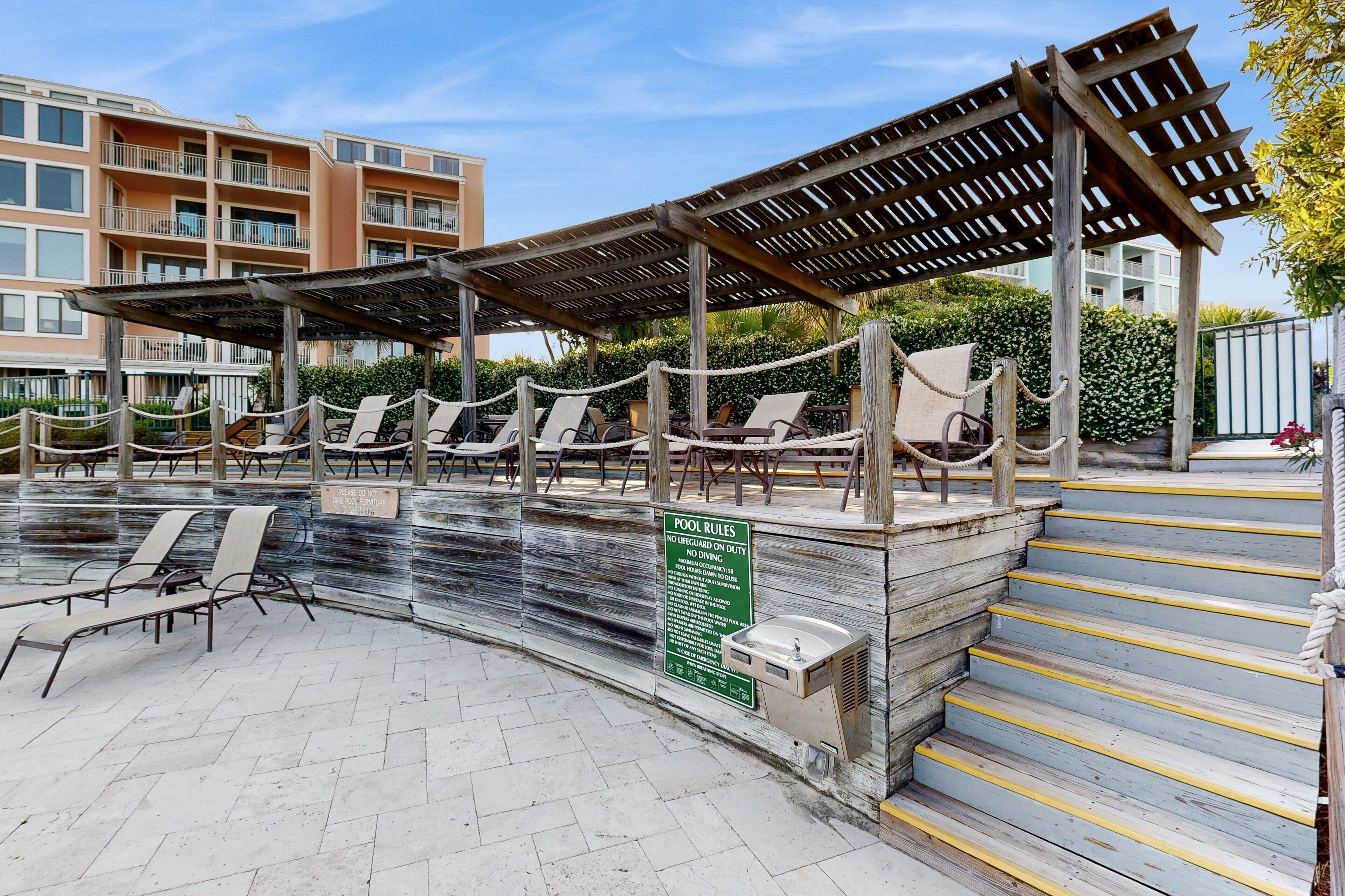 Zona de piscina con sillones de descanso, una pérgola de madera y un letrero con las normas de la piscina.