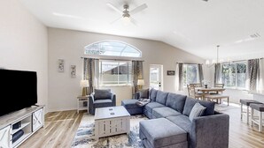 A spacious living room with a sectional sofa, a flat-screen TV, a ceiling fan, and a dining area with a table and chairs. - The Barefoot Bungalow House in Palm Coast With an Enclosed Pool & Grill (Palm Coast)