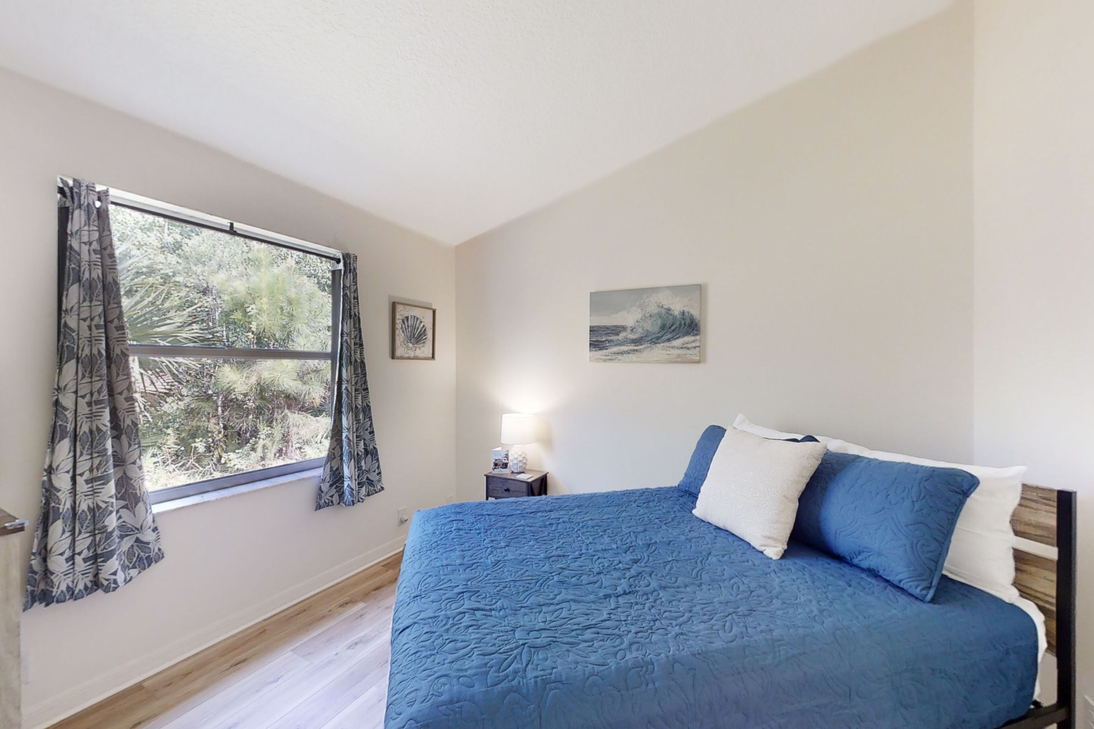 A bedroom with a large bed, a window with curtains, a nightstand with a lamp, and a framed picture on the wall.