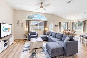 A modern living room with a sectional sofa, a coffee table, a TV, and a dining area with a table and chairs. - The Barefoot Bungalow House in Palm Coast With an Enclosed Pool & Grill (Palm Coast)