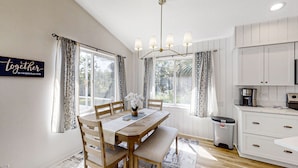 A dining area with a wooden table, chairs, and a bench. There is a chandelier above the table, and curtains on the windows. A trash can is visible near the table. - The Barefoot Bungalow House in Palm Coast With an Enclosed Pool & Grill (Palm Coast)
